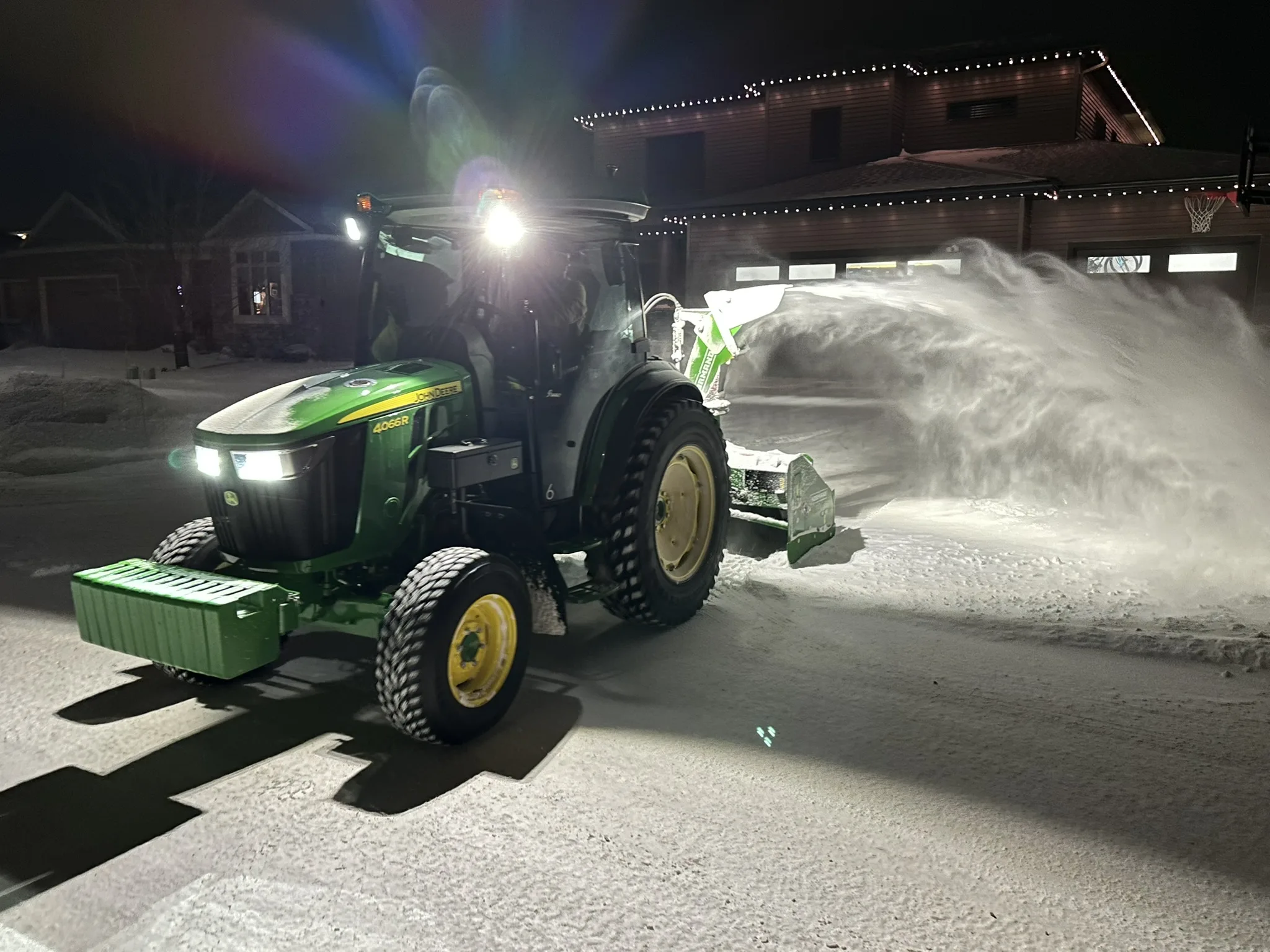 A Baker Snow & Ice tractor with inverted snowblower clearing a residential driveway